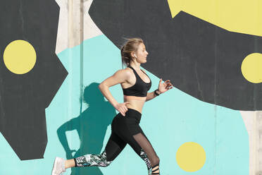 Young female athlete running by wall during sunny day - AMPF00194