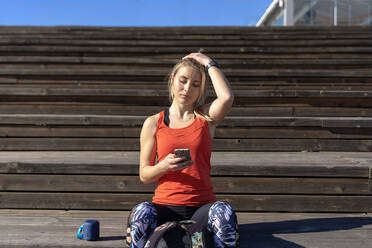 Junge Frau mit Kopf in der Hand, die ihr Smartphone benutzt, während sie an einem sonnigen Tag auf dem Pier sitzt - AMPF00200