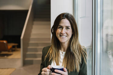 Smiling businesswoman with coffee mug leaning on glass window at office - XLGF01391