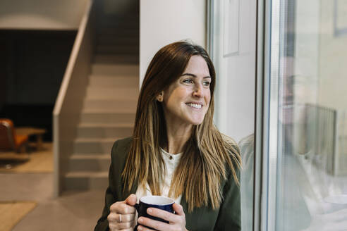 Nachdenkliche Geschäftsfrau mit Kaffeetasse, die durch ein Fenster im Büro schaut - XLGF01392