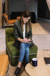 Female entrepreneur using smart phone while sitting on couch at office - XLGF01393