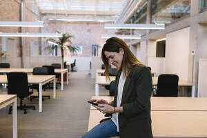 Geschäftsfrau, die ein Smartphone benutzt, während sie auf dem Schreibtisch im Büro sitzt - XLGF01400