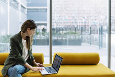 Unternehmerin mit Laptop an einer Glaswand, während sie auf dem Sofa im Büro sitzt - XLGF01406