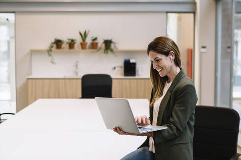 Lächelnde Unternehmerin mit In-Ear-Kopfhörern, die einen Laptop benutzt und sich auf einen Tisch im Büro stützt - XLGF01408