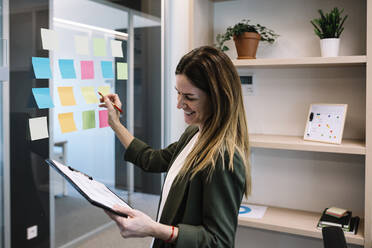 Smiling mid adult businesswoman holding clipboard while planning strategy on adhesive note at office - XLGF01419