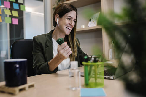 Cheerful female entrepreneur with in-ear headphones squeezing toy while working on laptop at office - XLGF01436