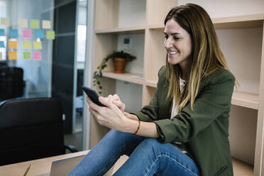 Smiling female entrepreneur using smart phone while sitting on desk at office - XLGF01443