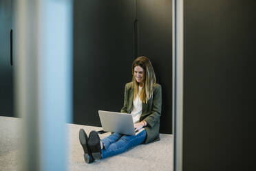 Smiling businesswoman using laptop while sitting on floor at office - XLGF01446