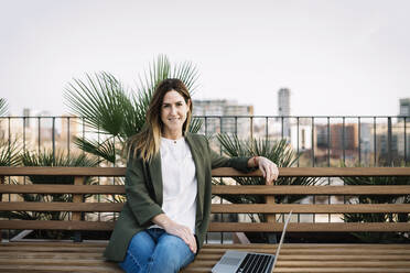 Businesswoman with laptop sitting on bench at building terrace - XLGF01450