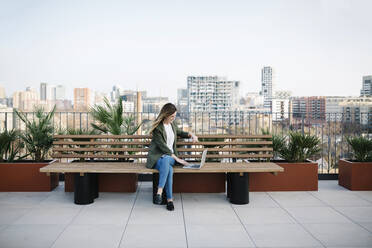 Frau, die einen Laptop benutzt, während sie auf einer Holzbank auf der Terrasse eines Gebäudes sitzt - XLGF01452