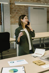Mittlere erwachsene Geschäftsfrau, die eine Kaffeetasse hält, während sie mit einem Smartphone im Büro telefoniert - XLGF01462