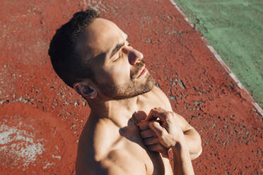 Shirtless man with hand on chest standing on sports court during sunny day - MIMFF00662