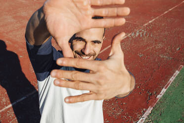 Lächelnder Mann schaut durch einen Fingerrahmen auf einem Sportplatz an einem sonnigen Tag - MIMFF00663