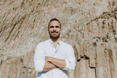 Smiling man with arms crossed standing in front of volcanic rock during sunny day - MIMFF00666
