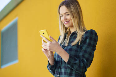 Lächelnde blonde Frau, die ein Mobiltelefon vor einer gelben Wand benutzt - JSMF02070