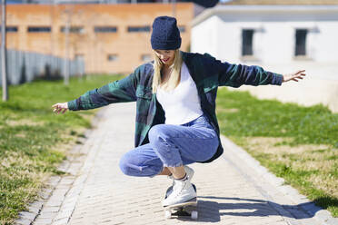 Lächelnde Frau hockt auf einem Skateboard an einem sonnigen Tag - JSMF02088