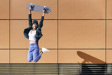 Unbekümmerte Frau, die ein Skateboard trägt, während sie vor eine Wand springt - JSMF02093