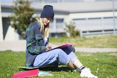 Junge Frau studiert auf dem Skateboard sitzend auf einem College-Campus an einem sonnigen Tag - JSMF02097