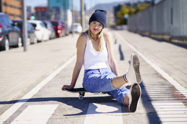 Blond woman sitting on skateboard during sunny day - JSMF02100