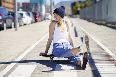 Smiling blond woman looking away while sitting on footpath during sunny day - JSMF02101