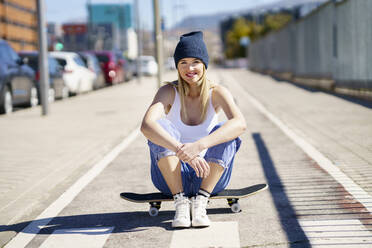 Lächelnde schöne Frau auf dem Skateboard sitzend an einem sonnigen Tag - JSMF02102