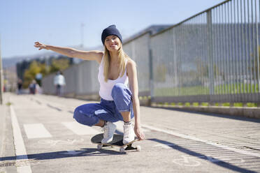 Lächelnde Frau mit ausgestreckten Armen auf dem Skateboard hockend an einem sonnigen Tag - JSMF02105