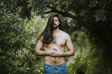 Mann ohne Hemd mit geschlossenen Augen steht und entspannt sich im Wald - MIMFF00671