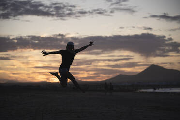 Aufgeregte Frau mit ausgestreckten Armen, die bei Sonnenuntergang am Strand springt - SNF01220