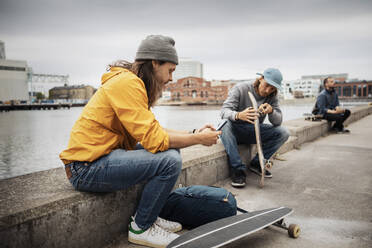 Männliche Freunde mit Smartphone und Skateboard auf einer Stützmauer sitzend - MASF22539