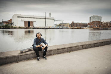 Lächelnder Mann, der ein Smartphone benutzt und auf einer Stützmauer am Kanal sitzt - MASF22543