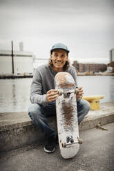 Portrait of smiling man with skateboard against canal - MASF22544