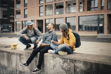 Glückliche männliche Freunde, die lachend auf einer Stützmauer vor einem Gebäude sitzen - MASF22549