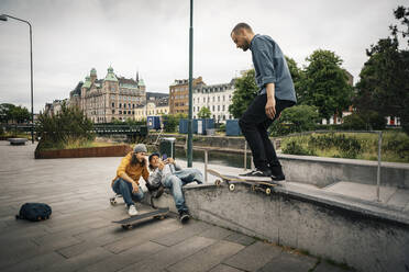 Freunde fotografieren mit dem Smartphone, während ein männlicher Skater mit einem Skateboard auf einer Stützmauer fährt - MASF22554