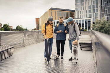 Freunde teilen ihr Smartphone mit männlichen Skatern, während sie auf einer Brücke stehen - MASF22565