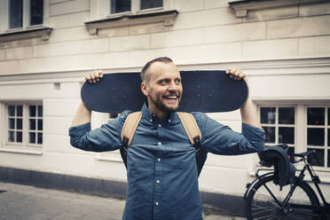Lächelnder Mann mit Skateboard, der wegschaut, während er auf dem Fußweg steht - MASF22570