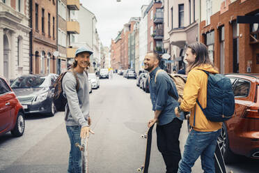 Glückliche Freunde unterhalten sich, während sie mit Skateboards auf der Straße in der Stadt stehen - MASF22577