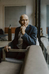 Portrait of male entrepreneur with hand on chin sitting over sofa at office - MASF22644