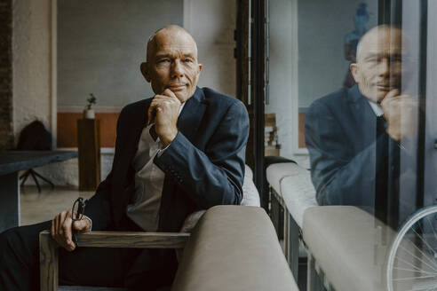 Portrait of businessman with hand on chin sitting over sofa at office - MASF22648