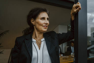 Smiling businesswoman contemplating while leaning on window at office - MASF22680