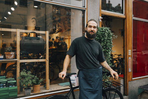 Porträt eines lächelnden männlichen Mechanikers auf einem Fahrrad vor einem Einzelhandelsgeschäft - MASF22728