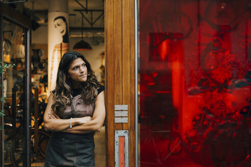 Weibliche Unternehmerin schaut weg, während sie an der Tür einer Werkstatt lehnt - MASF22733