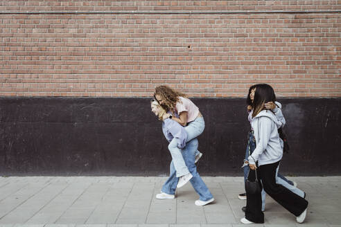 Glückliche Freundinnen tragen Huckepack, während sie auf dem Fußweg an der Mauer entlang gehen - MASF22793