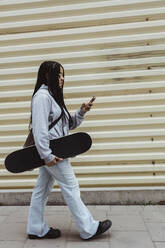 Teenager-Mädchen mit Skateboard, das sein Smartphone benutzt, während es auf dem Gehweg an einer Metallwand spazieren geht - MASF22794