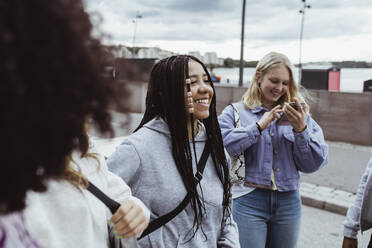 Teenager, der ein Smartphone benutzt, steht mit Freundinnen auf einem Fußweg - MASF22808