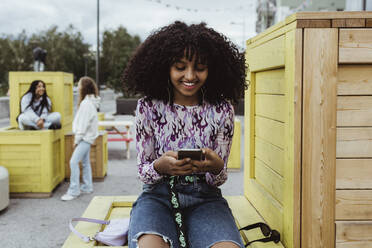 Lächelndes Teenager-Mädchen, das ein Smartphone benutzt, während es auf einer Kiste in der Stadt sitzt - MASF22822