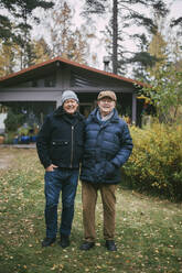 Portrait of smiling gay couple with hands in pockets standing in back yard - MASF22855