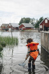 Rear view of boy wearing life jacket while standing in lake with fishing rod during summer - MASF22915