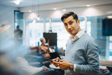 Porträt eines Geschäftsmannes mit Smartphone, während seine Kollegen im Hintergrund im Büro arbeiten - MASF22972