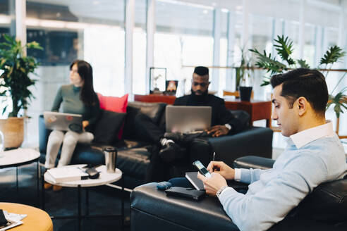 Männlicher Unternehmer, der sein Smartphone bei der Arbeit mit Kollegen im Büro benutzt - MASF22987