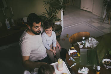 High angle view of father feeding sick son at table in living room - MASF23026